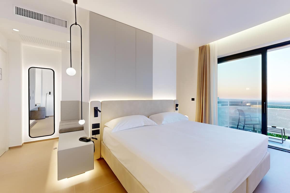 A modern hotel room with a large bed, a mirror, a bedside table, and a view of the city through a large window.