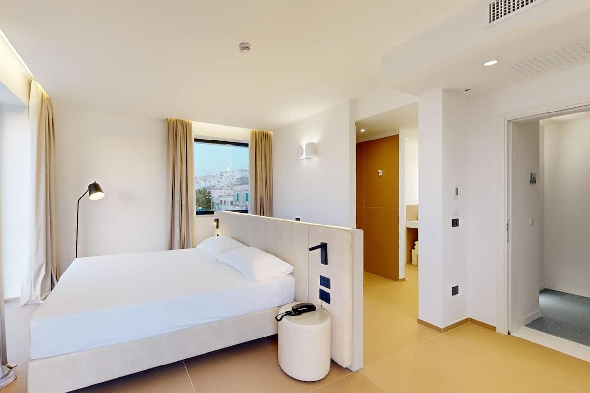 A modern hotel room with a bed, a bedside table, a lamp, and a view of the city through the window.