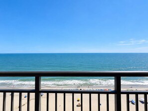 A beach view from a balcony with a railing, ocean, and beachgoers.