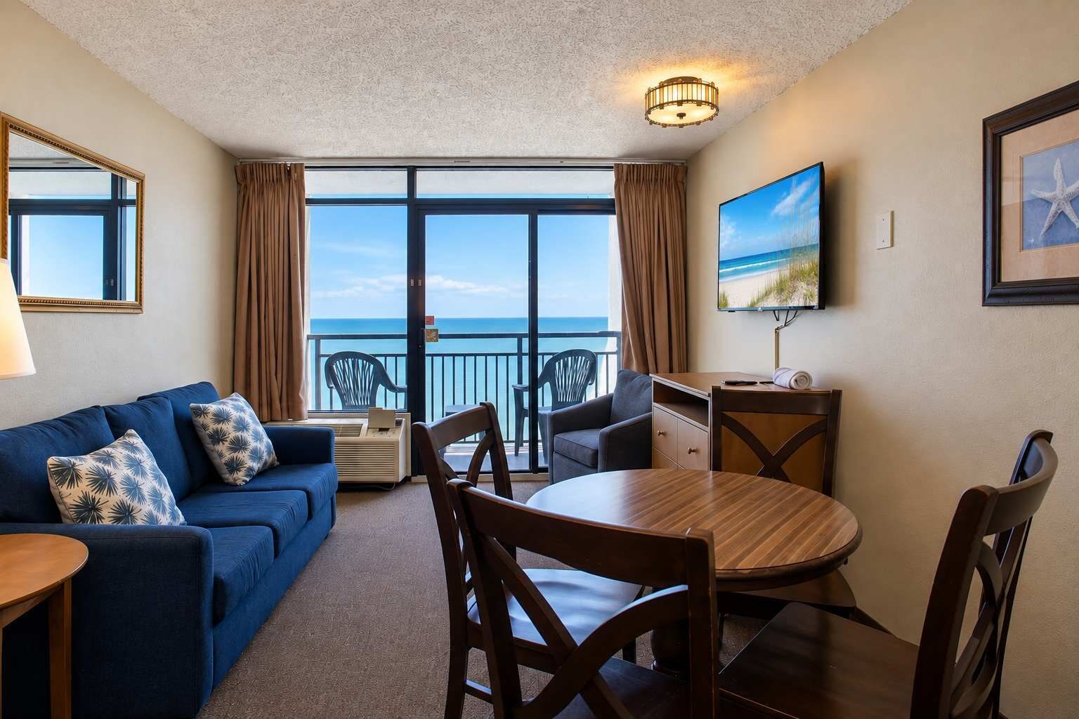 Ein Zimmer mit einem blauen Sofa, einem Holzesstisch und Stühlen. An der Wand ist ein Fernseher und ein Fenster mit Blick aufs Meer.