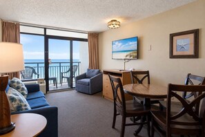 A hotel room with a balcony, a sofa, a dining table with chairs, a TV, and a framed picture on the wall.