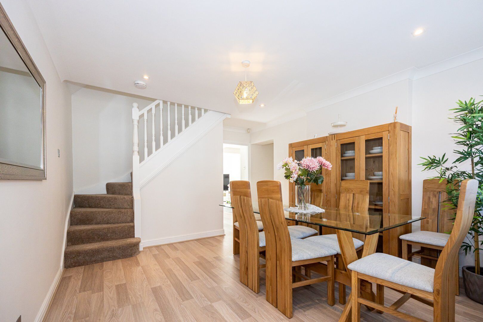A modern dining area with a glass table and wooden chairs, a staircase, and a wooden cabinet with shelves.
