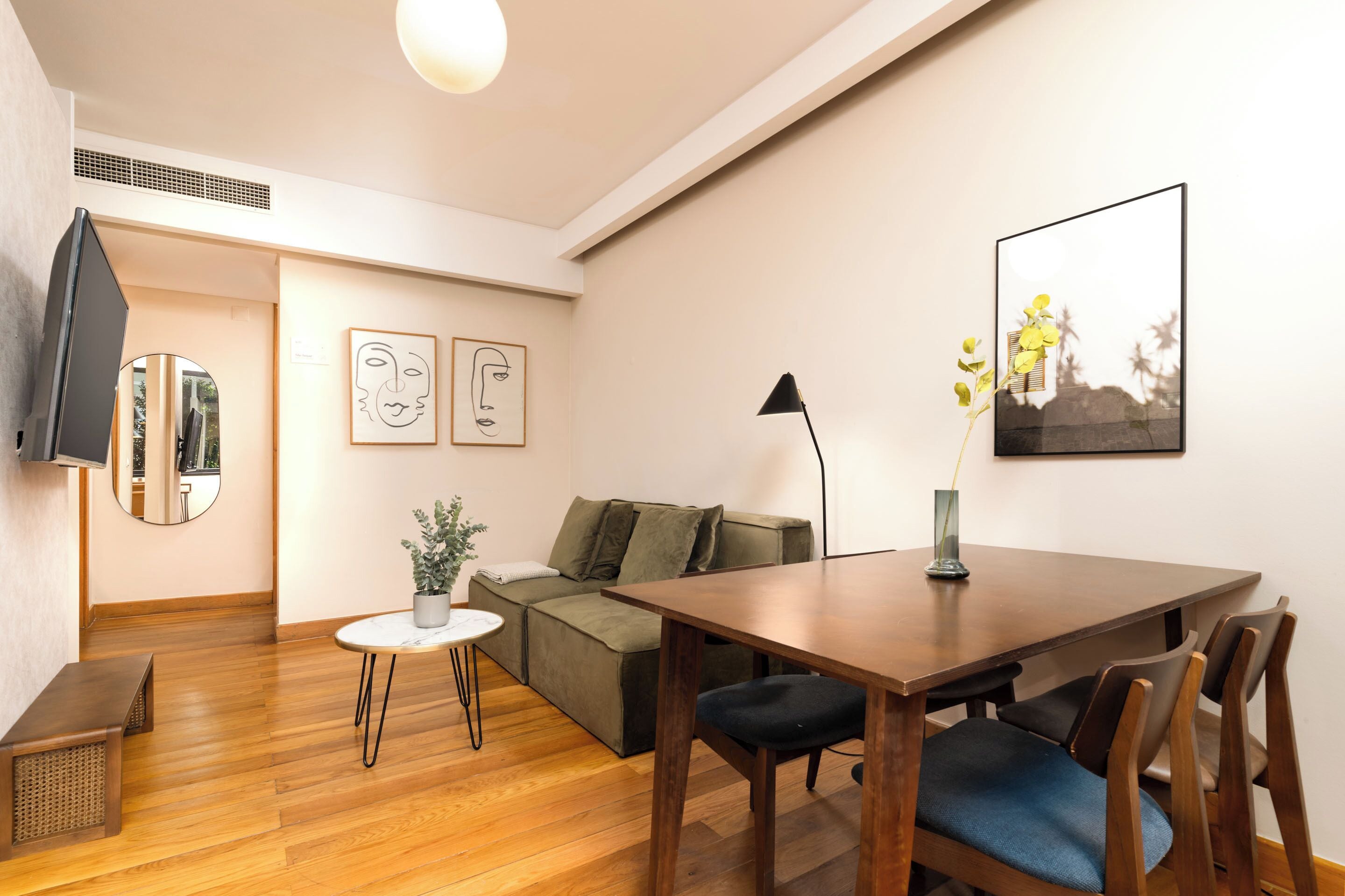 A modern living room with a wooden floor, a green sofa, a dining table with chairs, a small round table with a plant, and artwork on the walls.