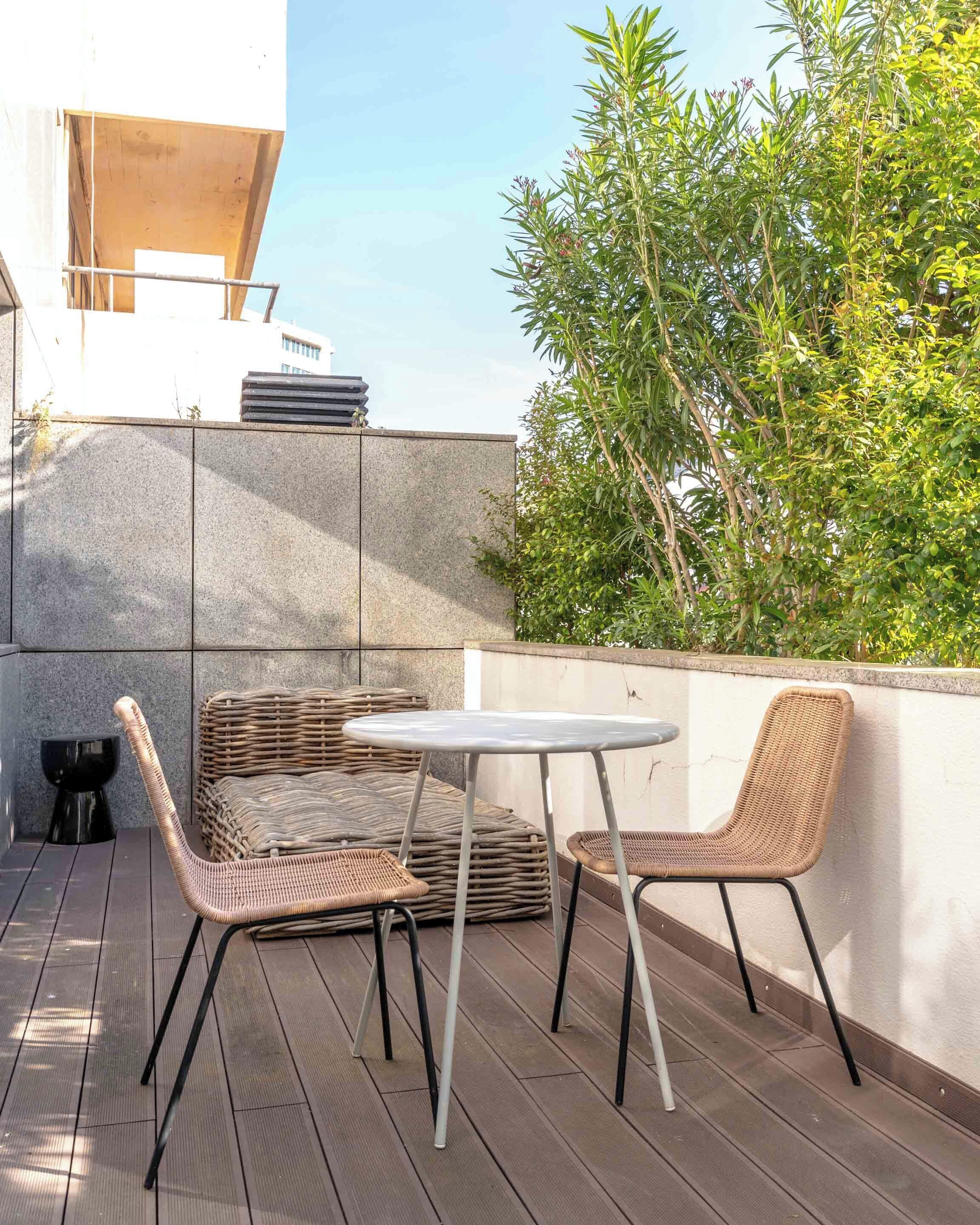 Terrassa exterior amb moble de vimet, taula rodona i vista a un edifici i verdura.