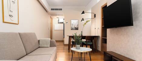 A modern living room with a sofa, a dining table with chairs, a TV mounted on the wall, and a potted plant.