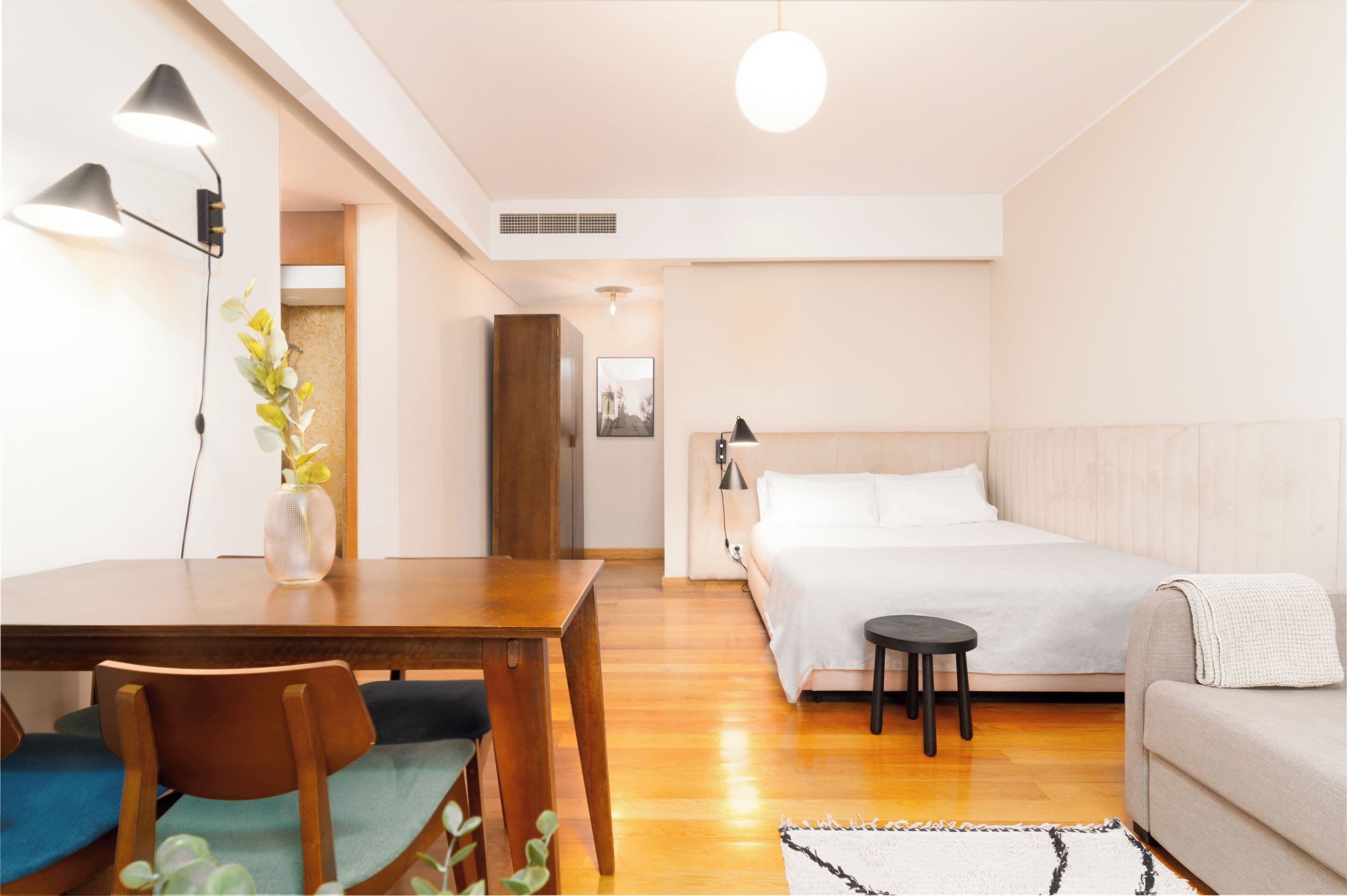 A modern bedroom with a wooden bed, a small bedside table, a sofa, a dining table with chairs, and a vase with flowers.
