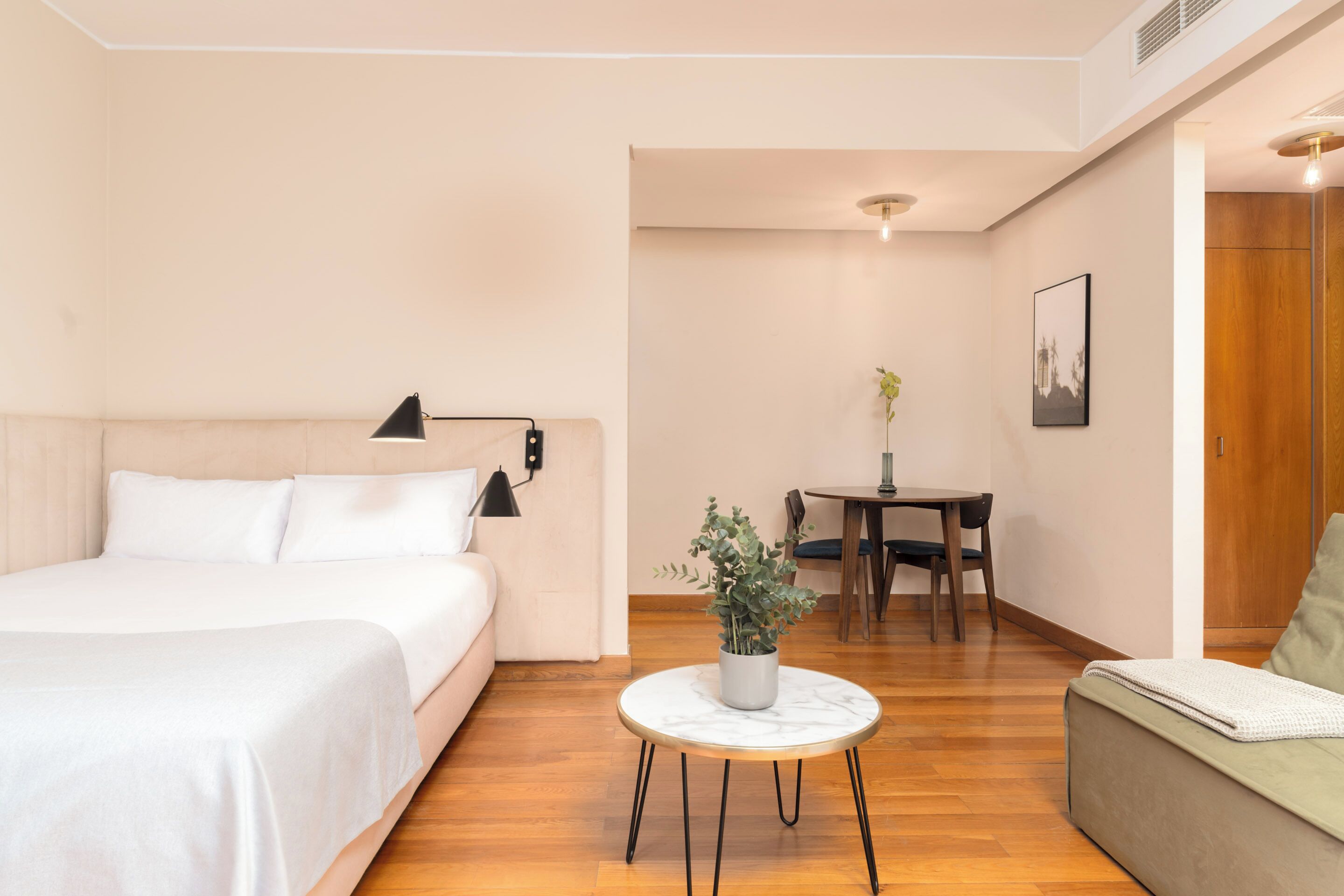 A modern hotel room with a bed, a round coffee table with a plant, a dining area with a table and chairs, and a wooden door.