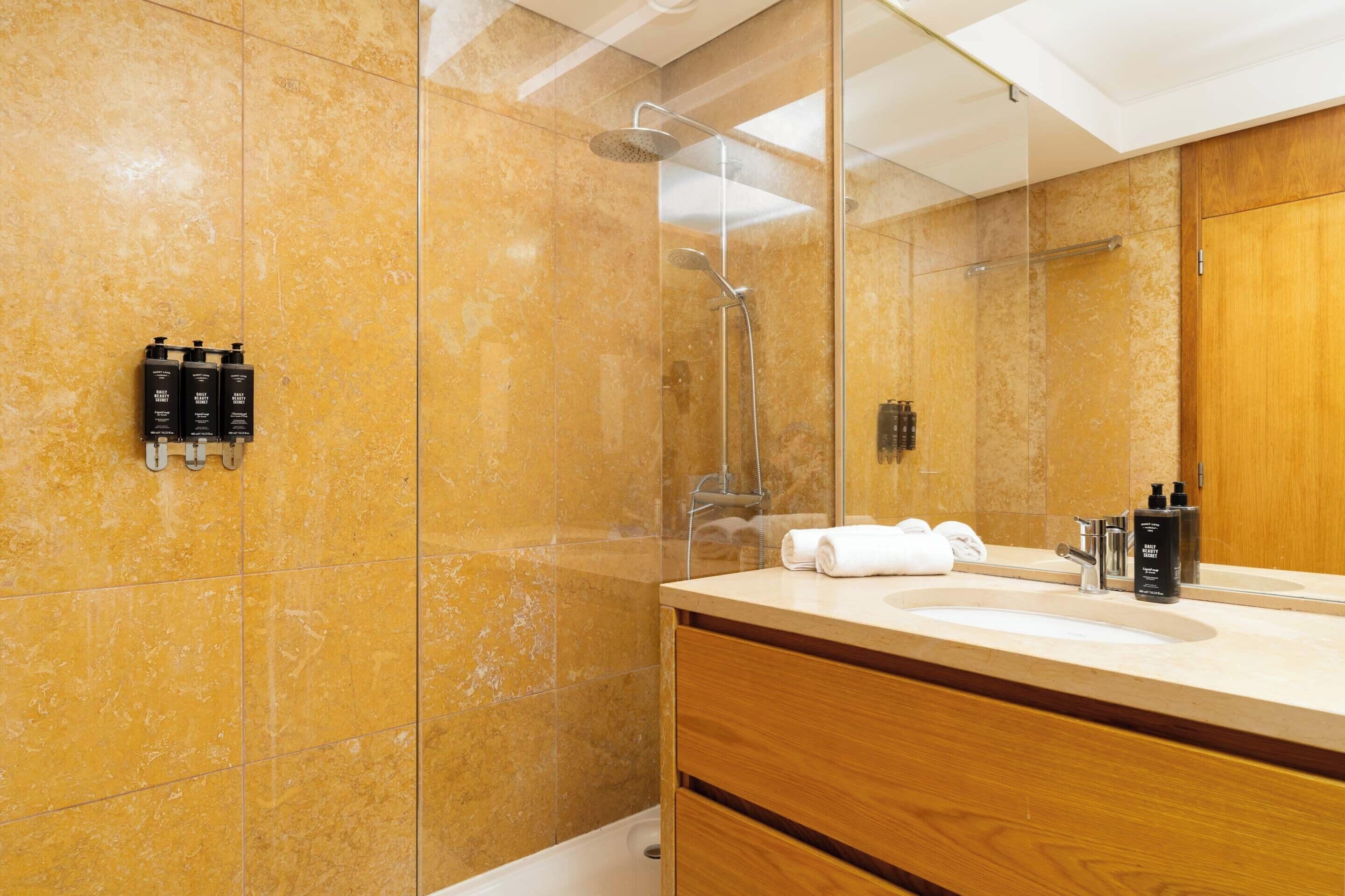 Un baño moderno con ducha envuelta en vidrio, tocador de madera con lavamanos y artículos de aseo en la encimera.