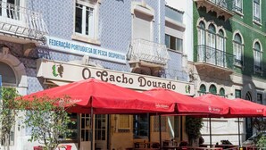 Ein Gebäude mit einer blau-weißen Fassade, einem Schild mit der Aufschrift 'O Cachoco Dourado' und Außenplätzen mit roten Sonnenschirmen.