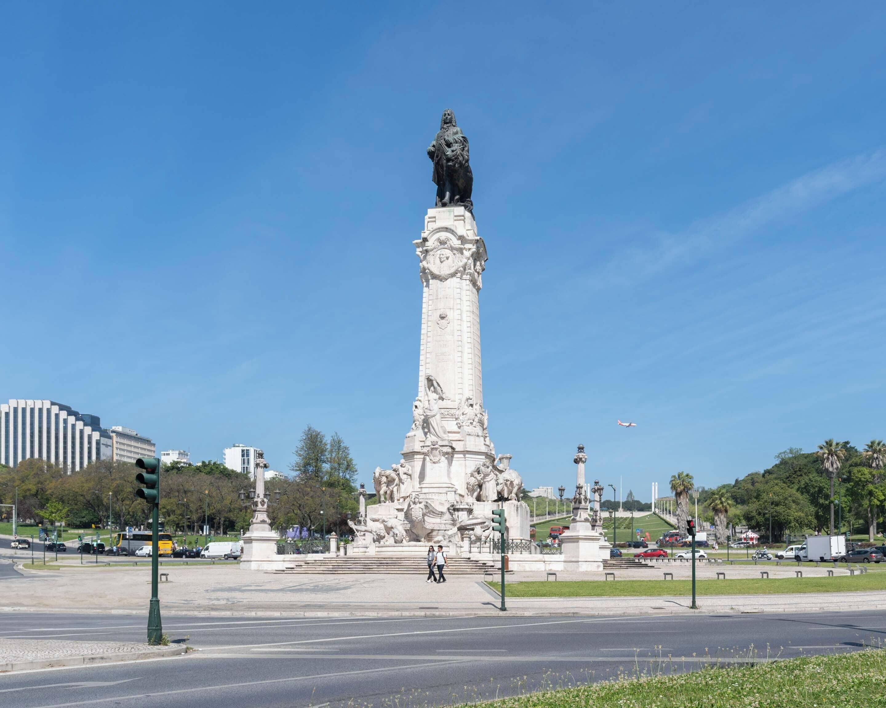 Ein großes, weißes Denkmal mit einer Statue auf der Spitze, umgeben von einem Platz mit Ampeln und einigen Fußgängern.