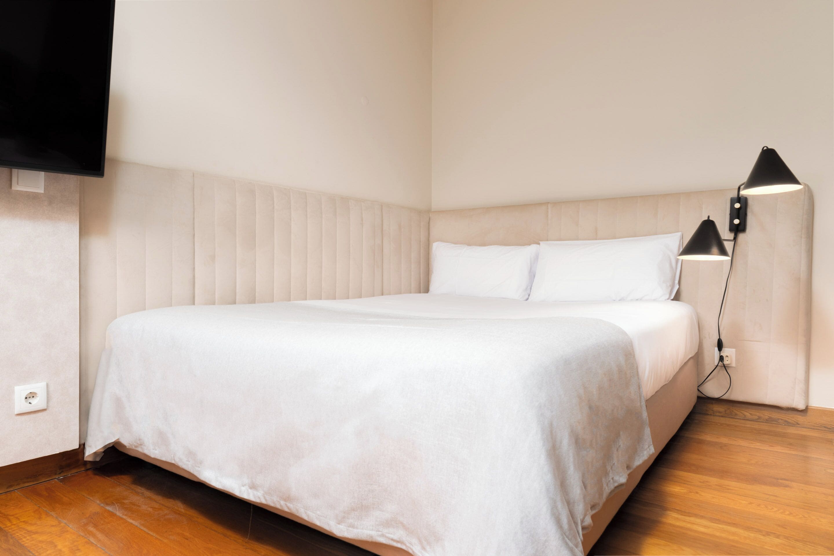 A neatly made bed with a white bedspread, a headboard with vertical slats, a black floor lamp, and a flat-screen TV mounted on the wall.