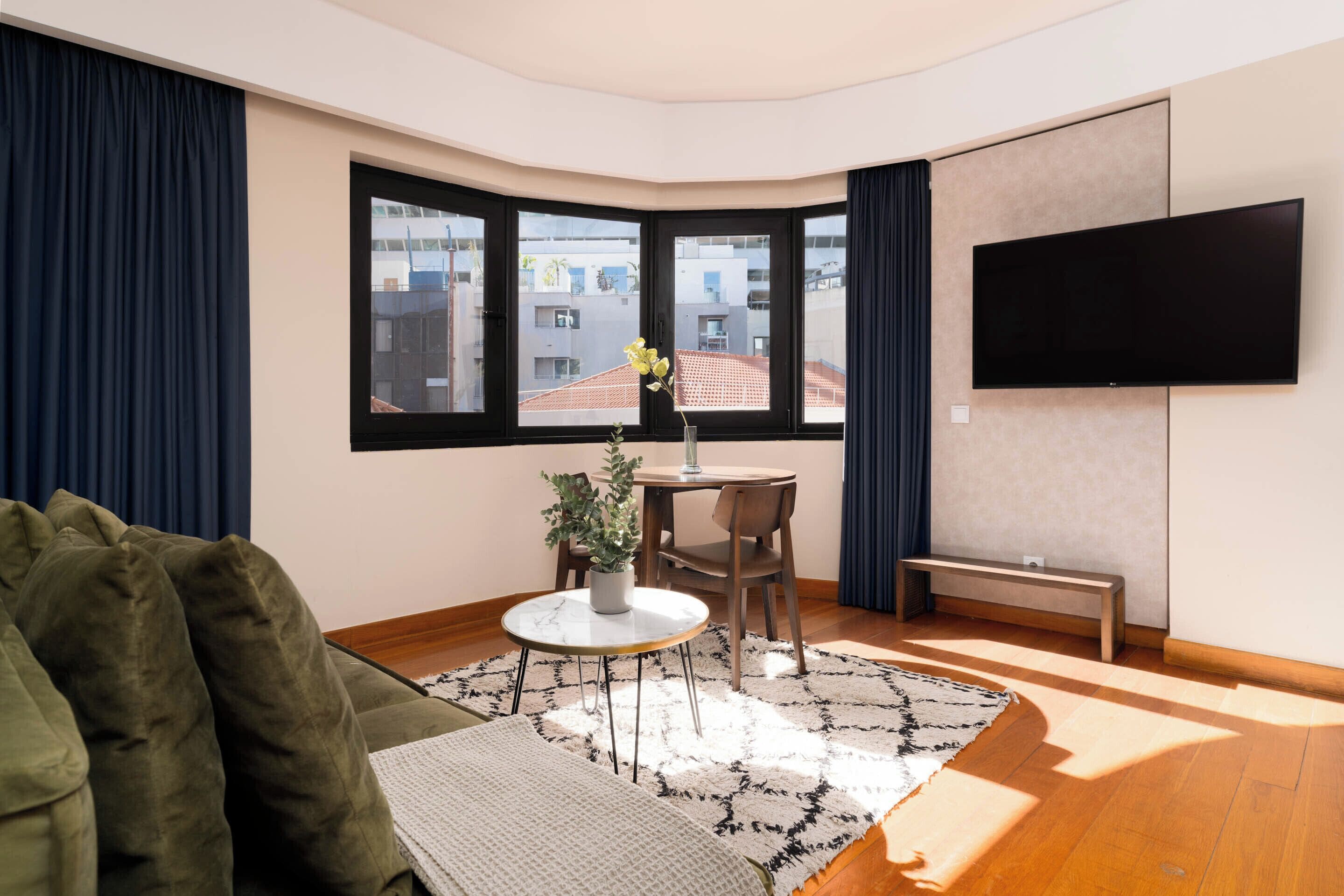 Een moderne woonkamer met een bank, een ronde tafel met stoelen, een potplant, een bankje en een grote televisie aan de muur.