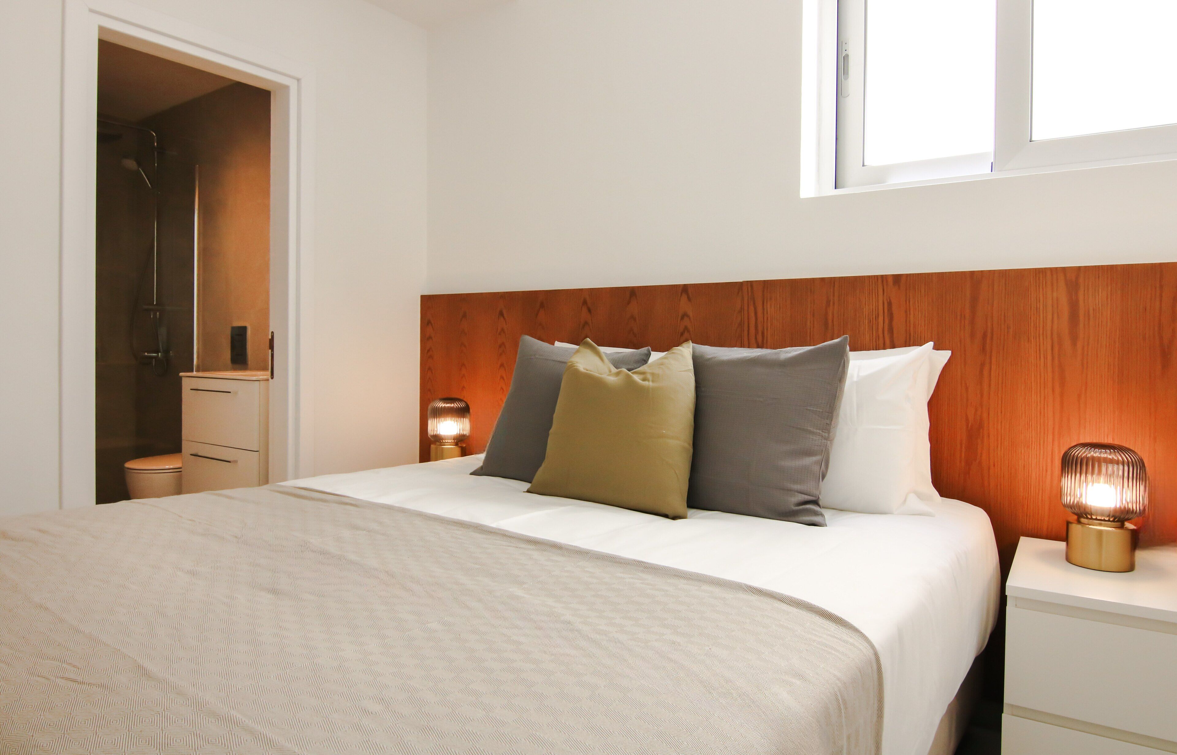A neatly made bed with a white comforter and pillows, a wooden headboard, a bedside lamp, and a small bedside table.