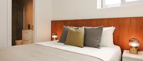 A neatly made bed with a white comforter and pillows, a wooden headboard, a bedside lamp, and a small bedside table.