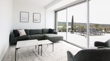 Una sala de estar moderna con un sofá seccional gris, una mesa de centro blanca y un amplio ventanal corredero que da a un espacio exterior con sillas de tomar el sol.