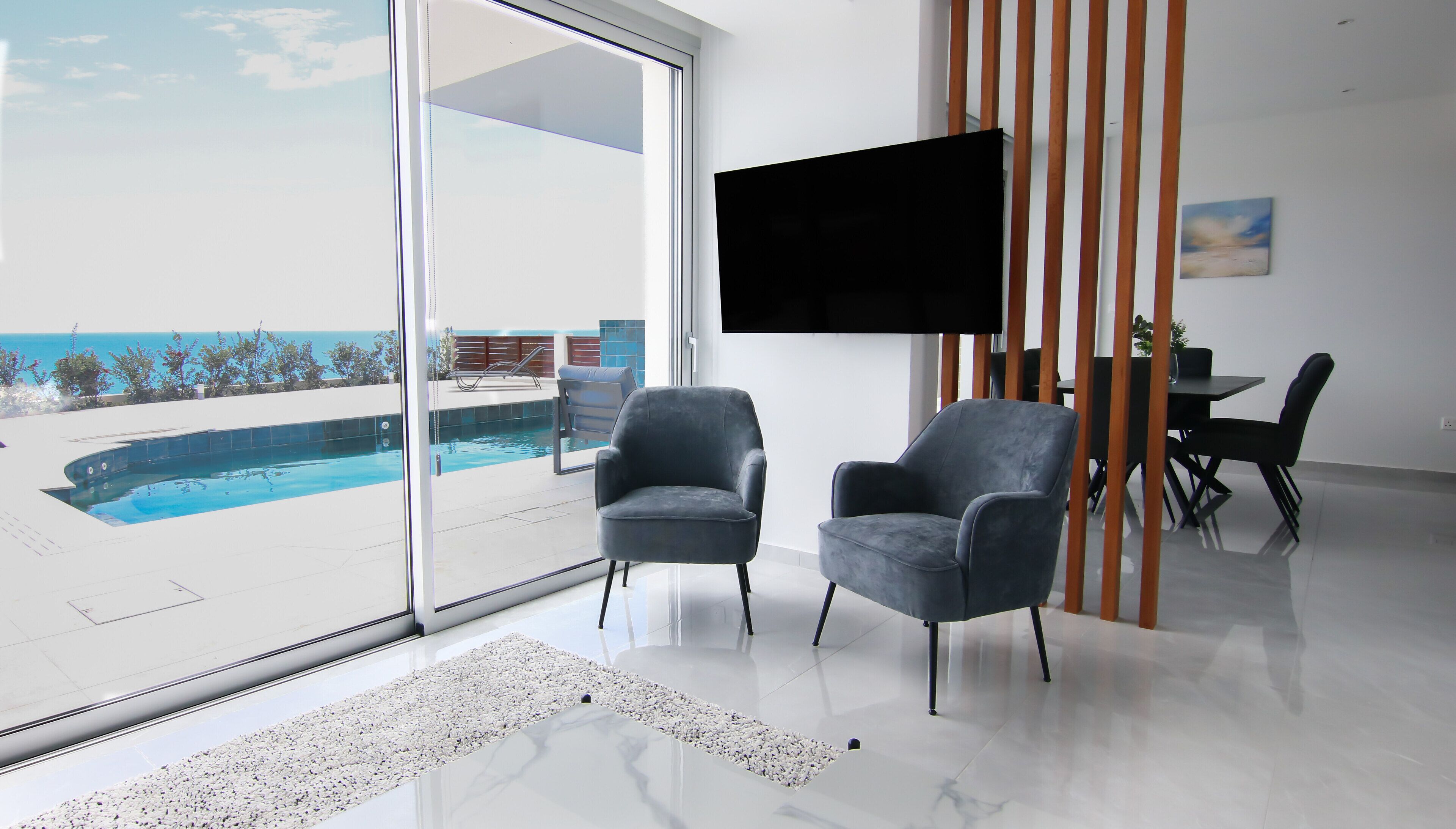 A modern living room with a large glass door leading to a pool, two grey armchairs, a flat-screen TV, and a dining area with a table and chairs.