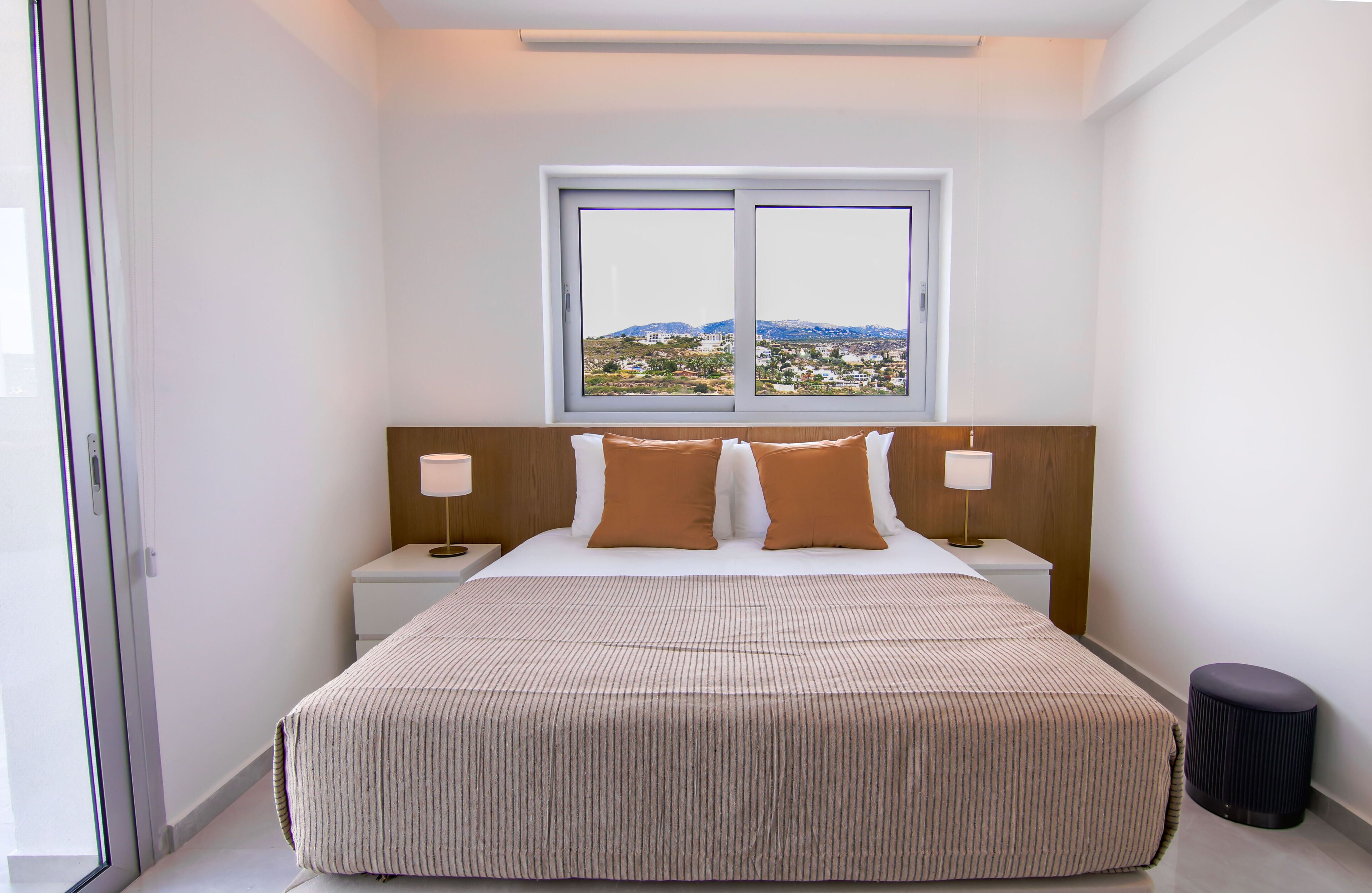A hotel room with a large bed, two bedside tables, a window with a view, and a small stool.