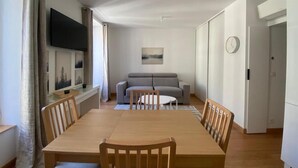 A modern living room with a wooden dining table, grey sofa, and a flat-screen TV mounted on the wall. - Détendez Vous Dans cet Appartement 2 Pièces Calme Élégant Rénové Entièrement (Nice)