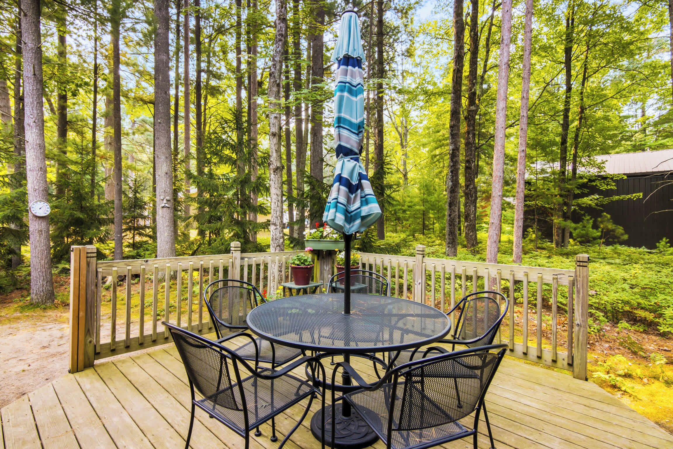Eine Holzterrasse mit rundem Tisch und Stühlen, umgeben von einem Hollerdgeschoss, in einer bewaldeten Gegend gelegen.