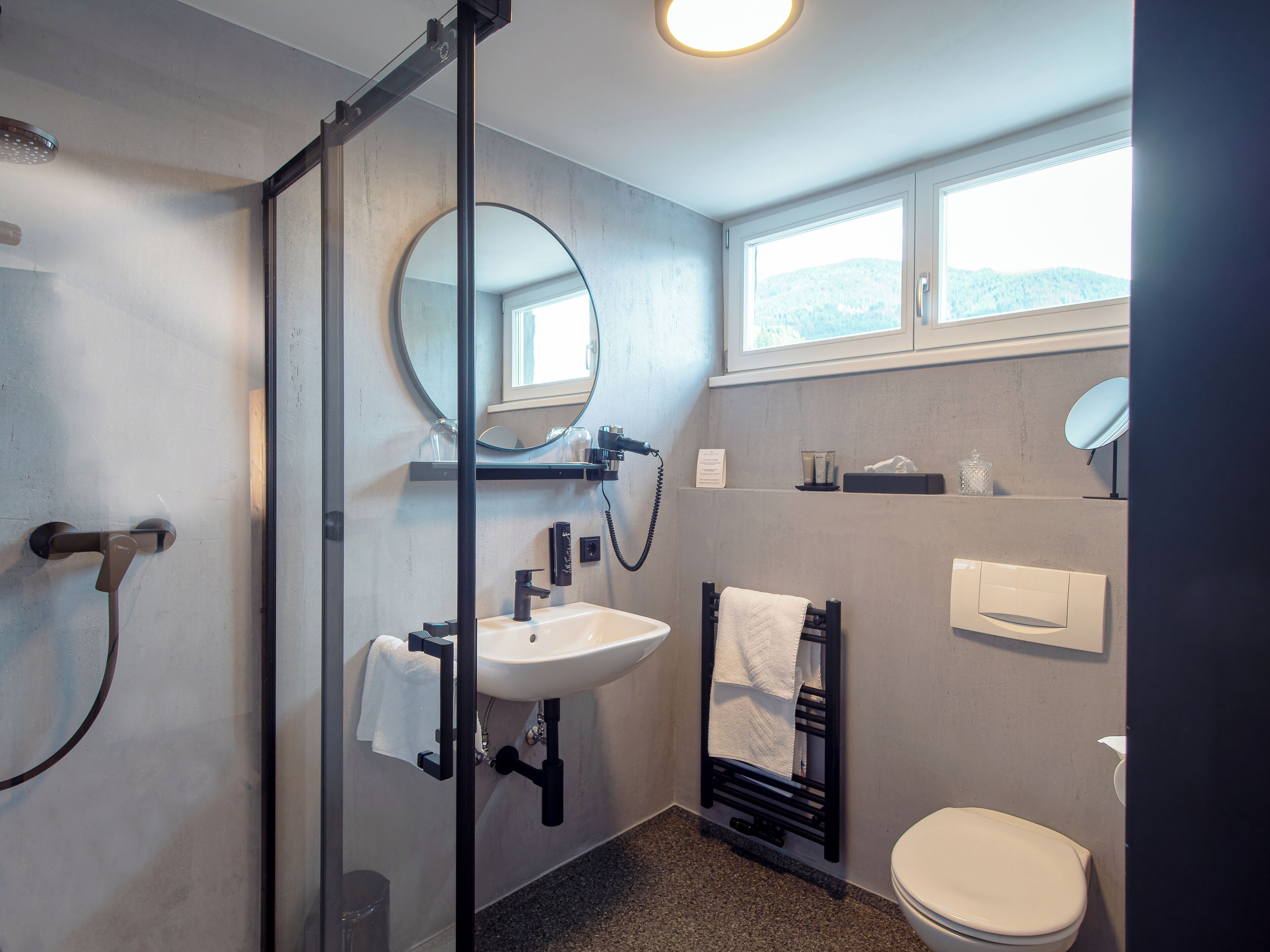 A modern bathroom with a glass shower enclosure, a sink with a mirror above it, a toilet, and a window providing natural light.