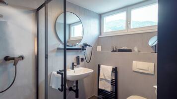 A modern bathroom with a glass shower enclosure, a sink with a mirror above it, a toilet, and a window providing natural light.