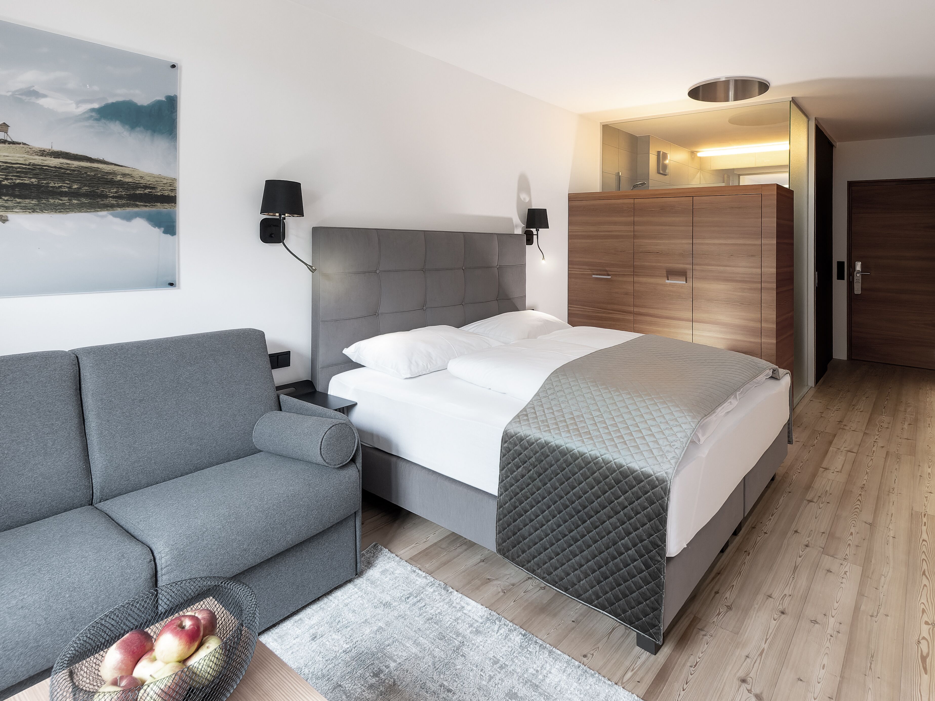 Un dormitorio moderno con un sofá gris, una cama con edredón acolchado, un armario de madera y una lámpara fijada a la pared.