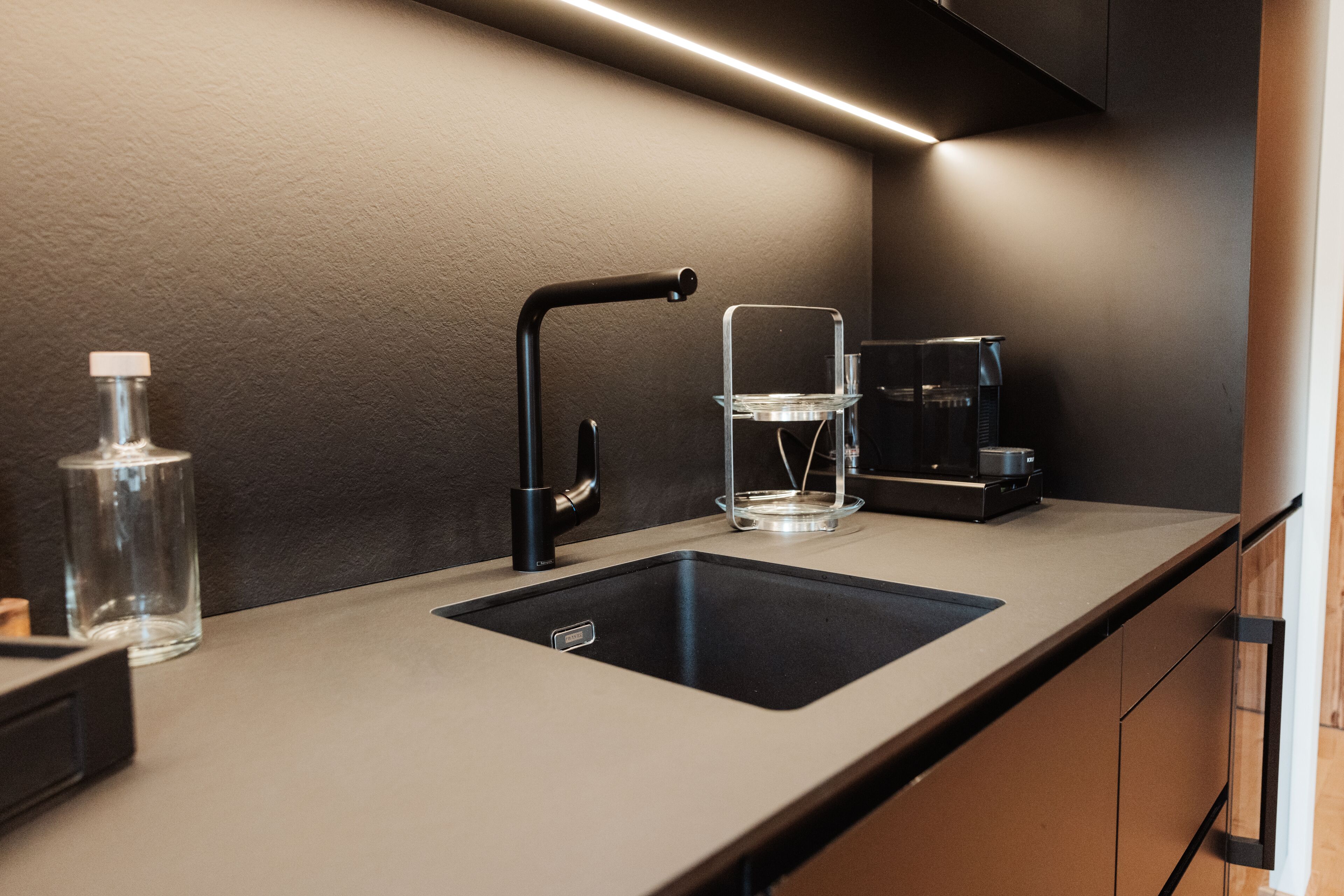 Een moderne keuken met een zwarte spoelbak, een koffieapparaat en een glazen fles.
