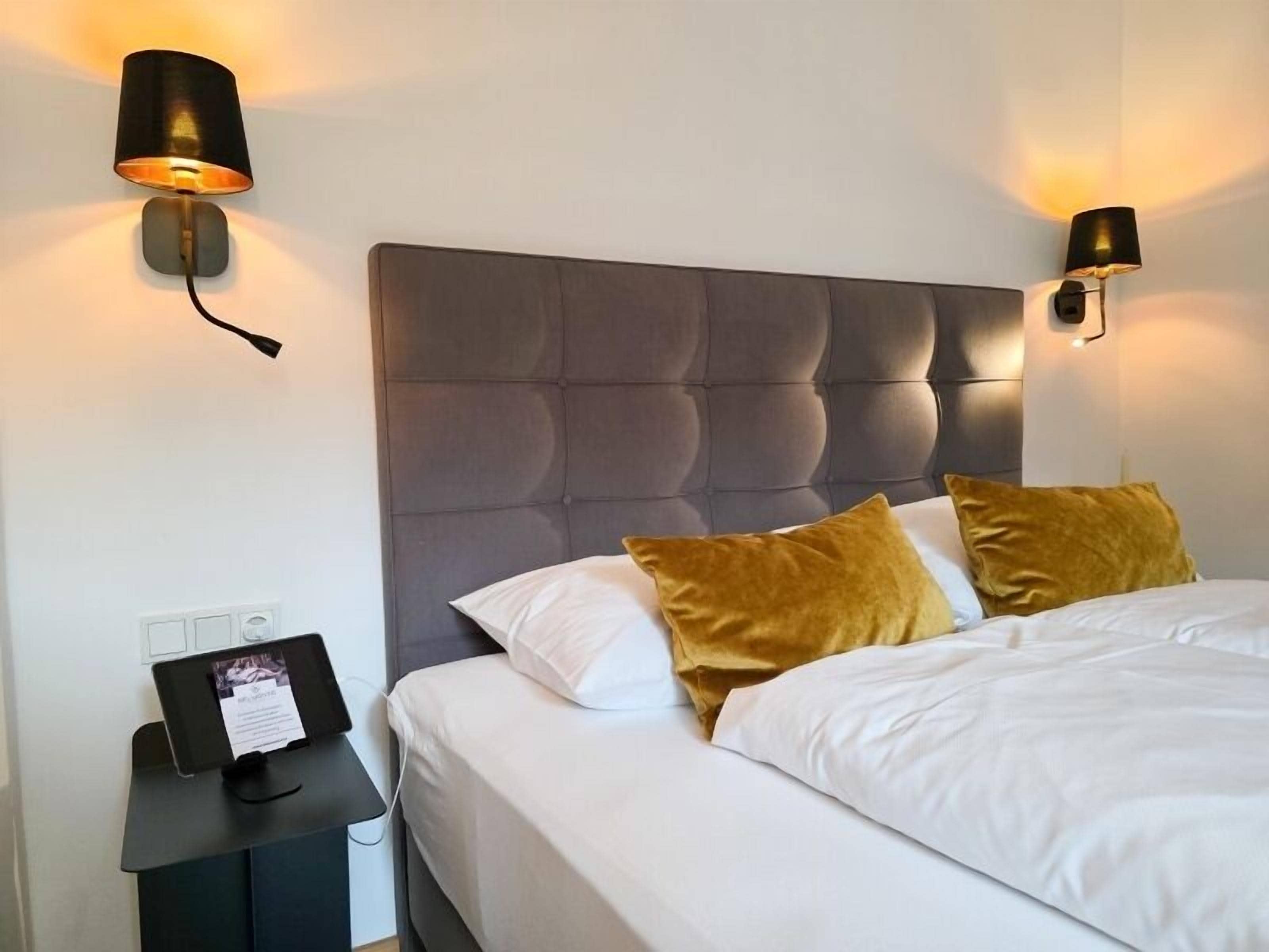 A hotel room with a large bed, two wall-mounted lamps, a bedside table with a photo frame, and a power outlet.
