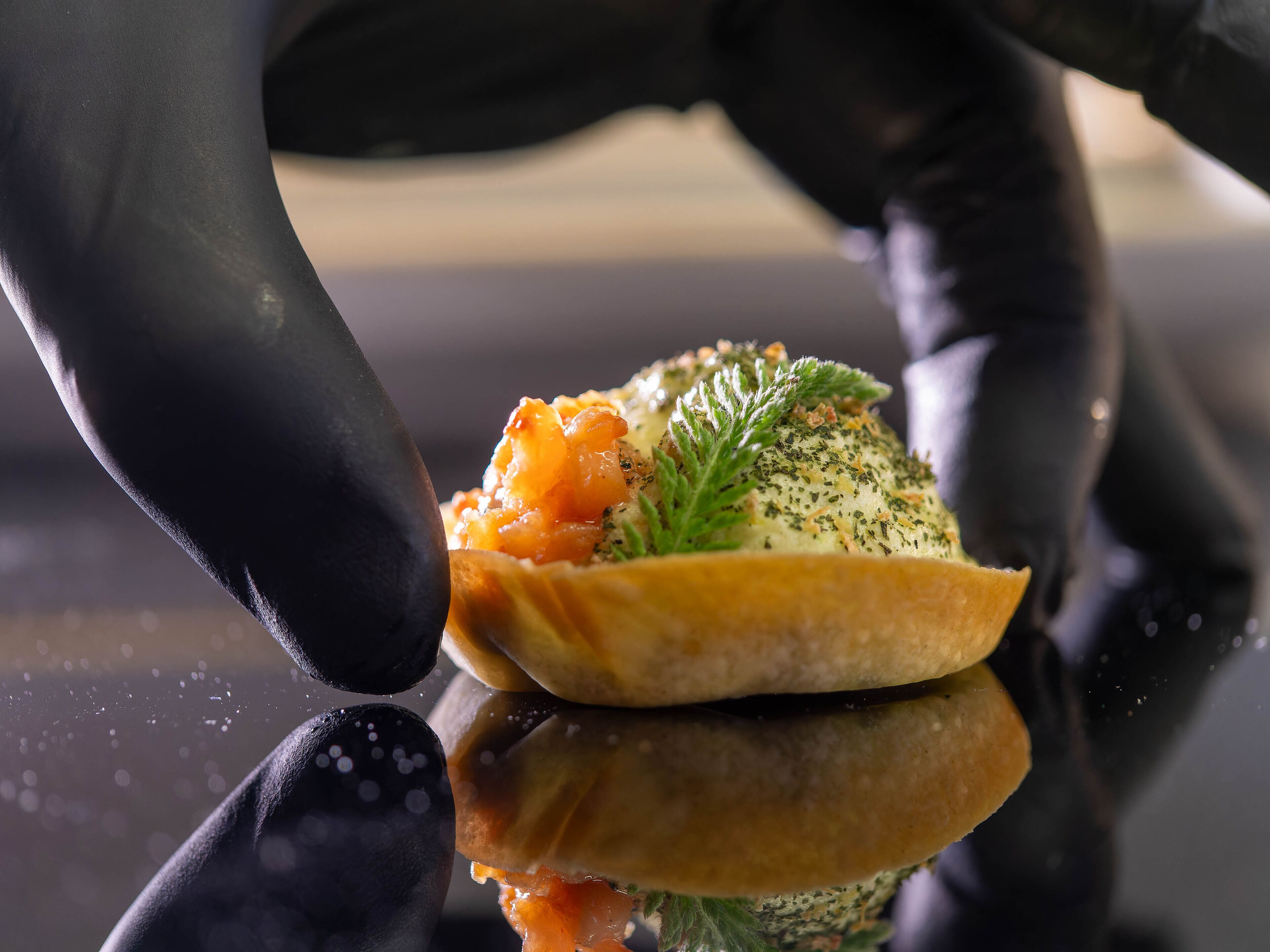 A close-up of a person wearing black gloves handling a dish with a creamy sauce and garnished with herbs.