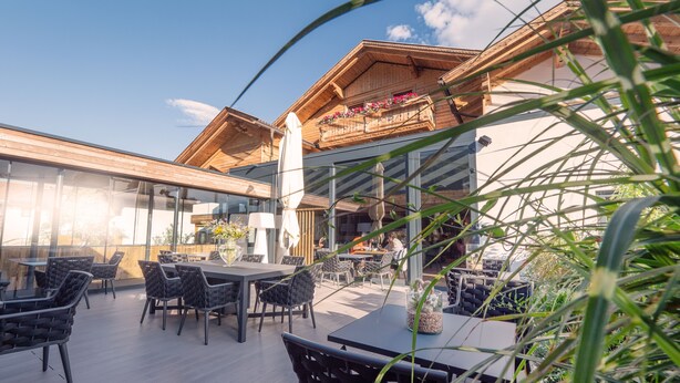 Een terras buiten met houten tafels en stoelen, een glazen overkapping en uitzicht op het interieur van het gebouw.