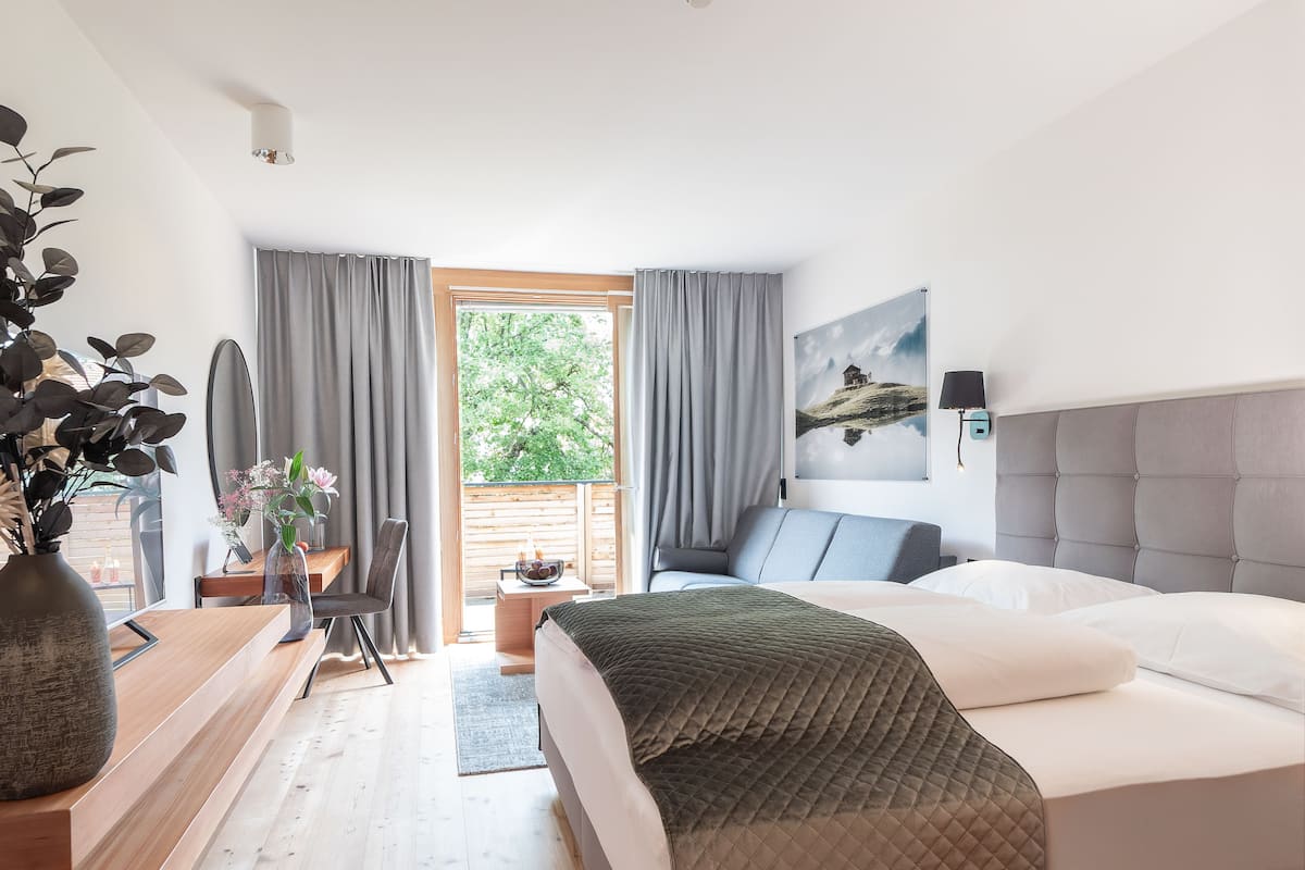 Habitación de hotel con cama, escritorio con silla, ventana con cortinas, una planta y un cuadro en la pared.