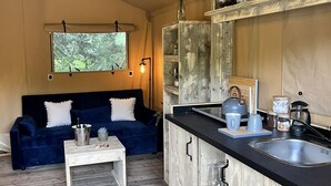 A compact living space with a kitchenette, a dark blue sofa, a small white table, and a window with a view of greenery. - Kingfisher Safari Tent (Droitwich)