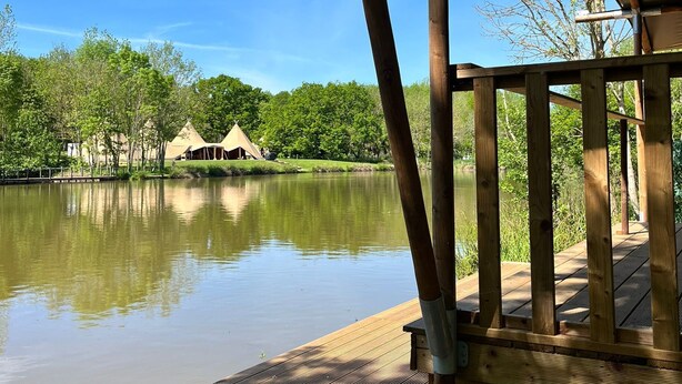 A wooden deck overlooking a calm lake with a thatched-roof structure in the background. - Kingfisher Safari Tent (Droitwich)