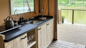 A wooden cabin with a kitchen featuring a sink, kettle, and glassware. A deck overlooks a body of water.