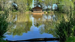 A wooden cabin with a deck, surrounded by a pond and trees, under a clear blue sky.