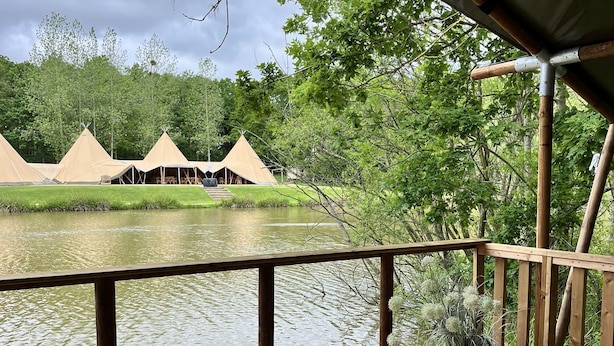A wooden deck with a table set for dining, overlooking a calm lake and several conical tents in the background.