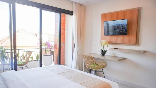 Quarto de hotel com cama, escrivaninha, cadeira, televisão presa à parede e varanda com cadeiras e plantas.
