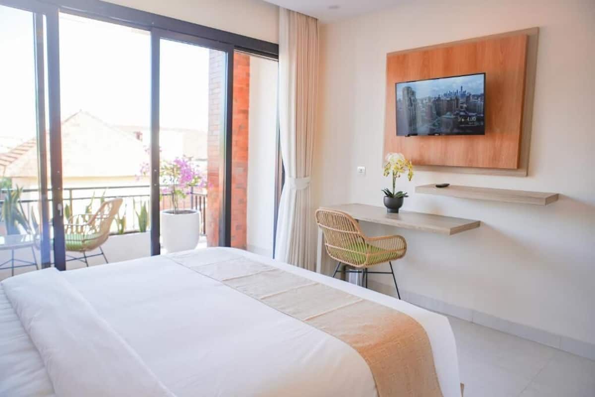 Une chambre d’hôtel comprenant un lit, un bureau, une chaise, un téléviseur fixé au mur et un balcon avec des chaises et des plantes.