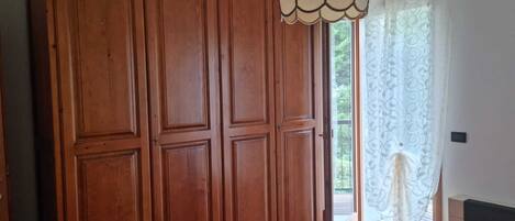 A bedroom with a wooden wardrobe, a bed with a red and white checkered bedspread, and a window with patterned curtains.