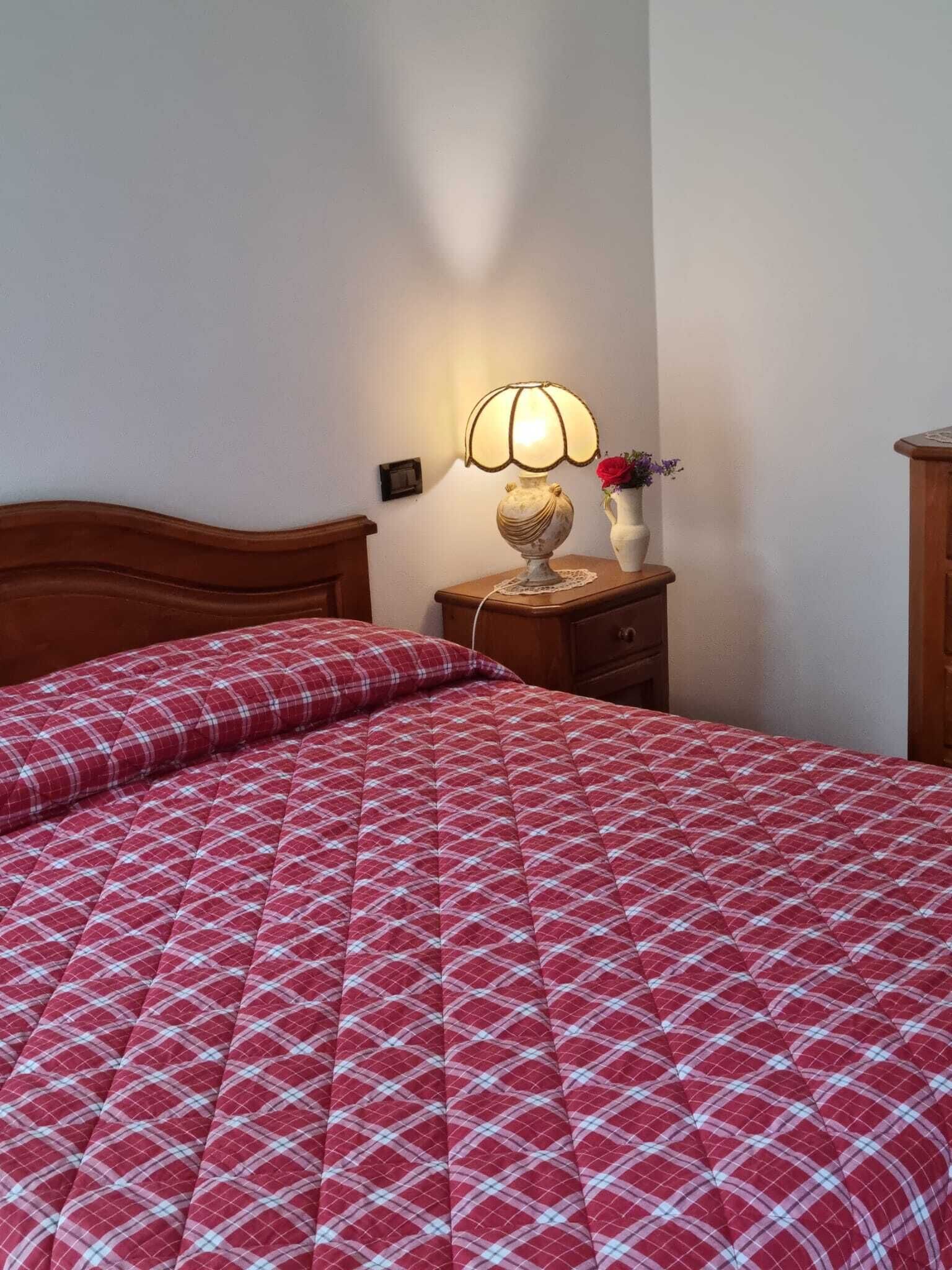 Una cama bien tendida con una colcha roja y blanca a cuadros, un cabecero de madera, una mesita de noche con una lámpara y un florero con flores.