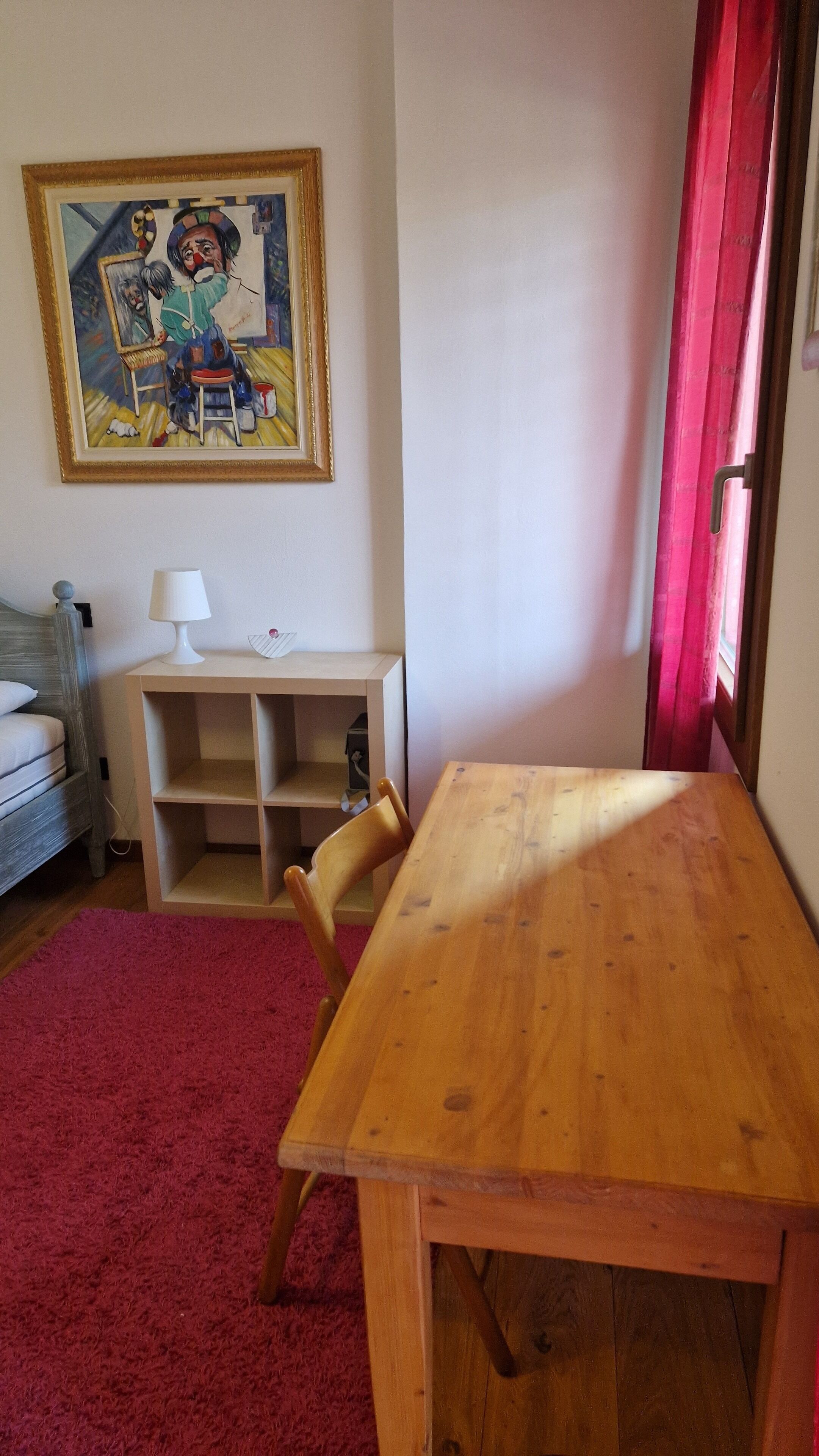 Een kamer met een houten tafel, een rood vloerkleed, een schilderij aan de muur en een raam met rode gordijnen.