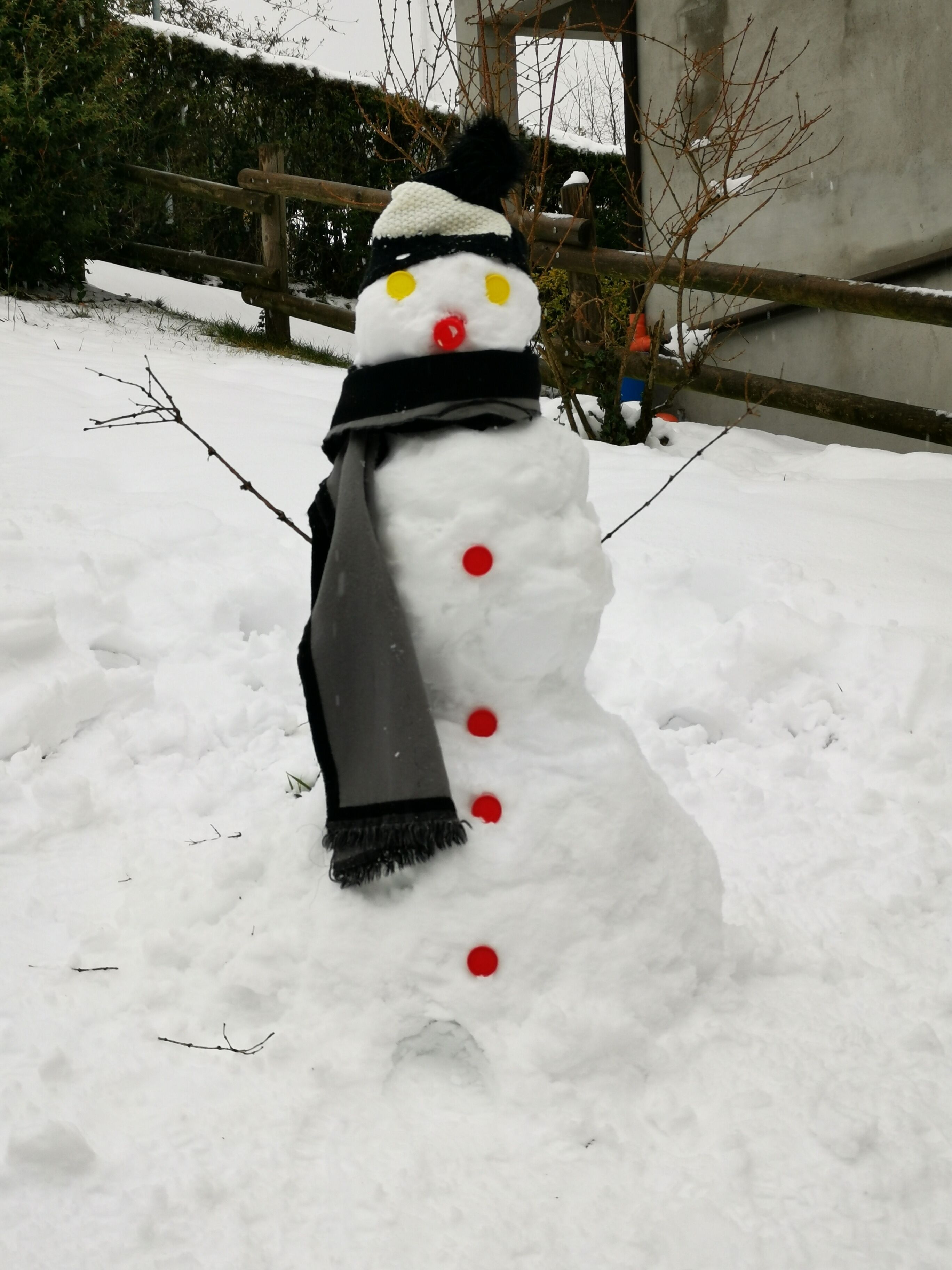 Un muñeco de nieve con bufanda negra y sombrero, de pie en un espacio exterior nevado.