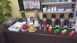 A breakfast setup with various food items and containers on a table, a shelf with dishes, and a window with curtains. - Hotel American (Ariquemes)