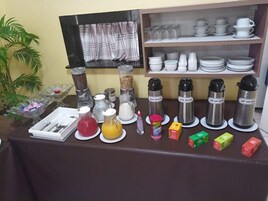 Un desayuno dispuesto con diferentes alimentos y recipientes sobre una mesa, una estantería con vajilla y una ventana con cortinas.