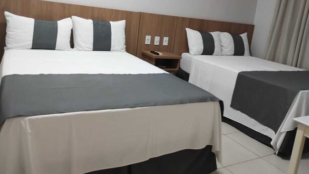 A hotel room with two beds, each with a grey and white bedspread, a wooden headboard, and a small bedside table with a lamp. - Hotel American (Ariquemes)