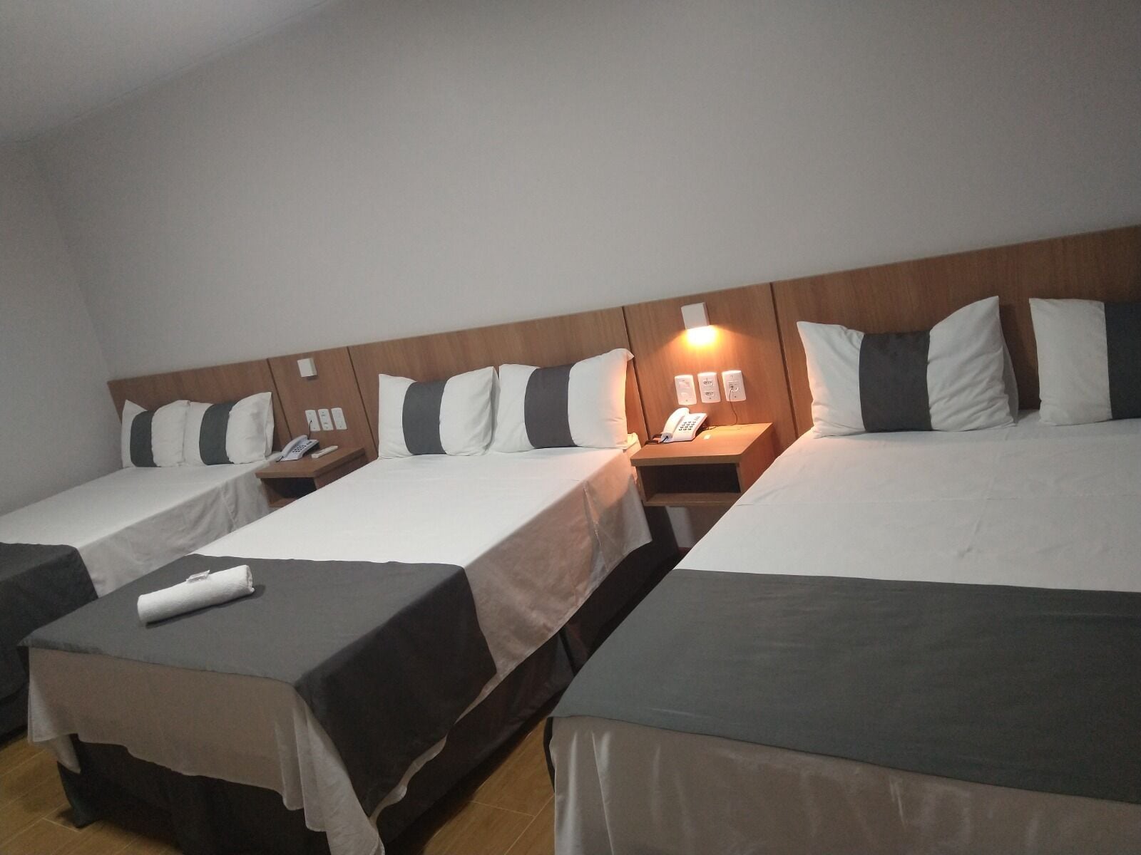 A hotel room with two single beds and a larger bed, each with a wooden headboard and white bedding. There are bedside tables with lamps and a phone on one of them.