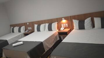 A hotel room with two single beds and a larger bed, each with a wooden headboard and white bedding. There are bedside tables with lamps and a phone on one of them.