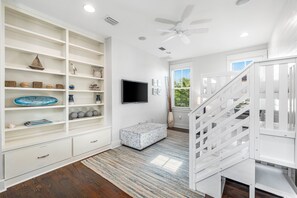 A spacious room with a white staircase, a flat-screen TV mounted on the wall, and a large white shelving unit filled with decorative items. - Indulge in Luxury at WaterColor – Pet-Friendly, Electric Cart, Steps to Pool & Close to Seaside (Santa Rosa Beach)