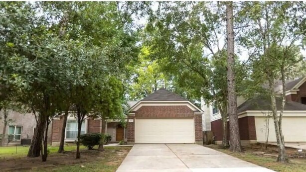 A residential driveway leading to a garage with a brick house, surrounded by trees and a grassy lawn.