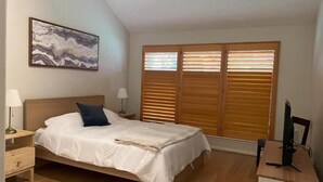 A bedroom with a bed, nightstands, a desk, and a large window with blinds.